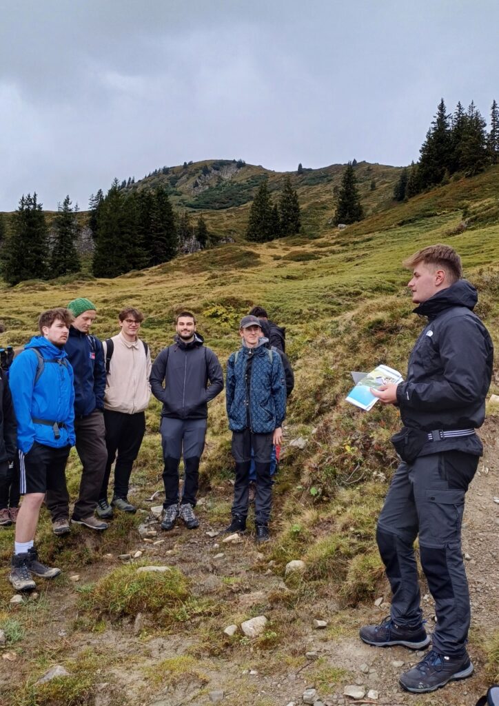 8 Schüler in hügeliger Landschaft mit Tannen im Hintergrund. 1 Schüler packt eine Wanderkarte aus.