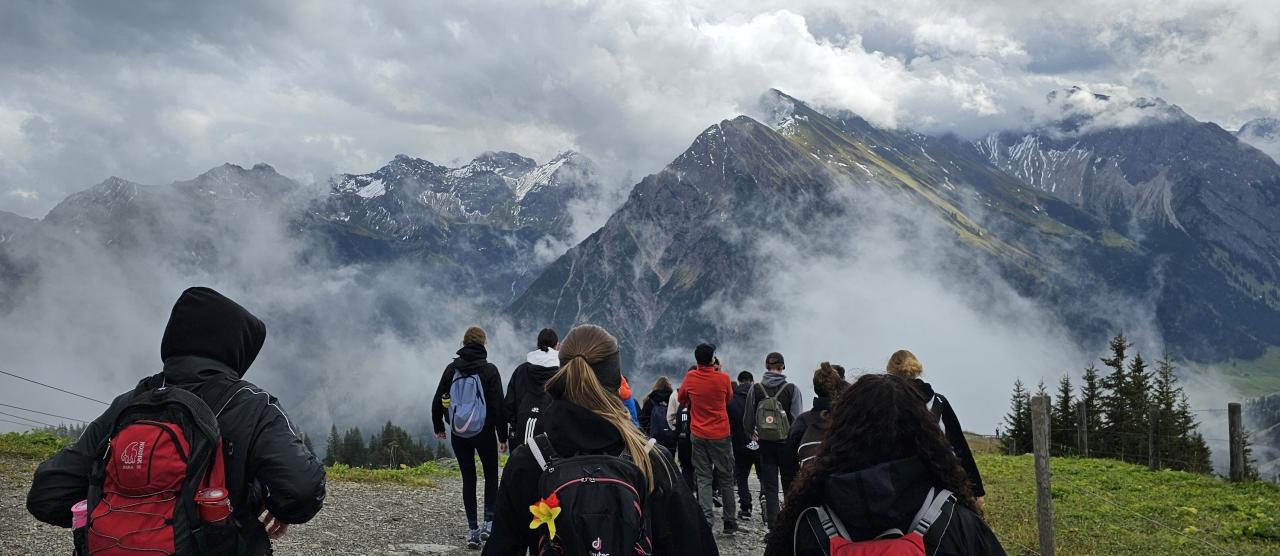 Schüler wandern Weg entlang mit Blick auf die Spitzen der Alpen. Nebel steigt von den Seiten auf.