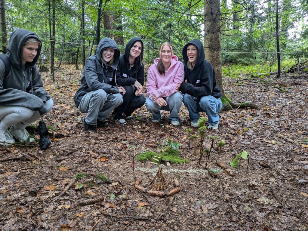 5 Schülerinnen im Wald, die in der Hocke um ihre Kunstwerke aus Tannenzapfen, Ästen und Moos sitzen und lachen