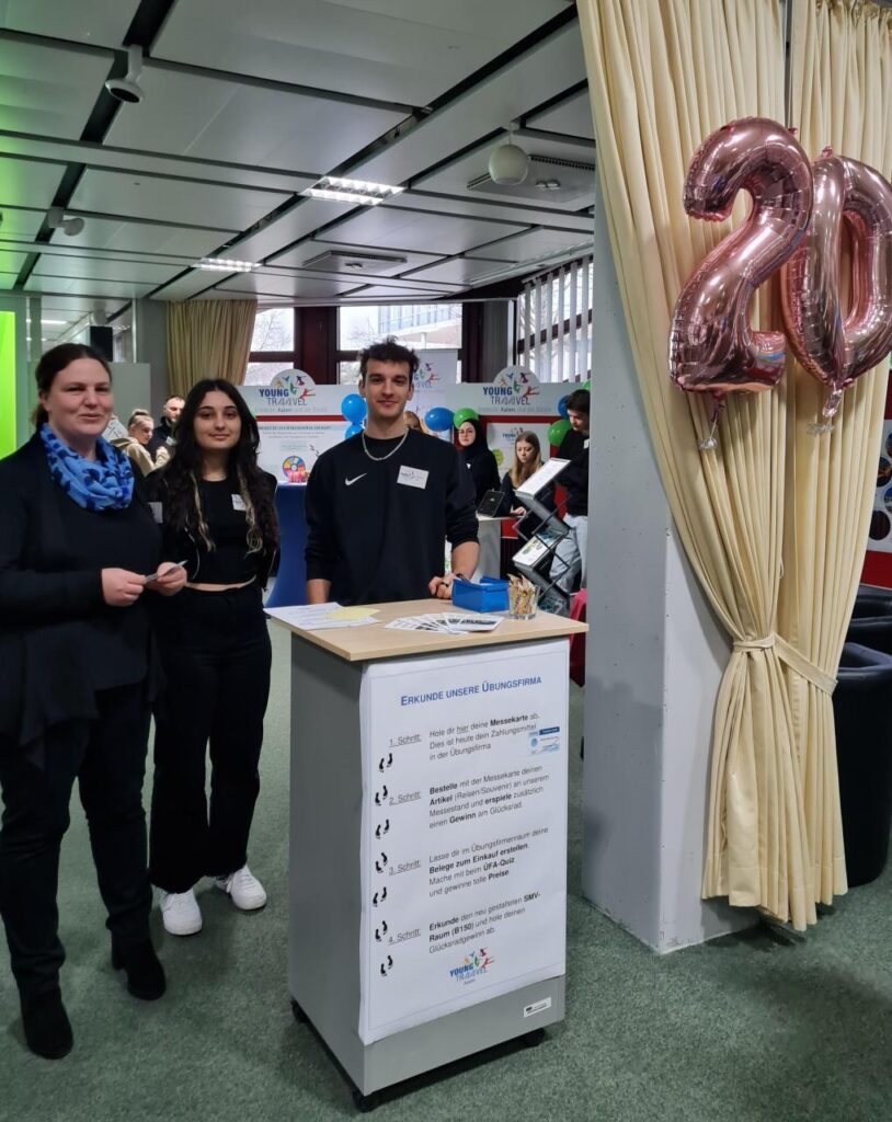 2 Schüler stehen am Empfang der Übungsfirma. Im Vordergrund hängt ein rosa Luftballon mit der Zahl 20.