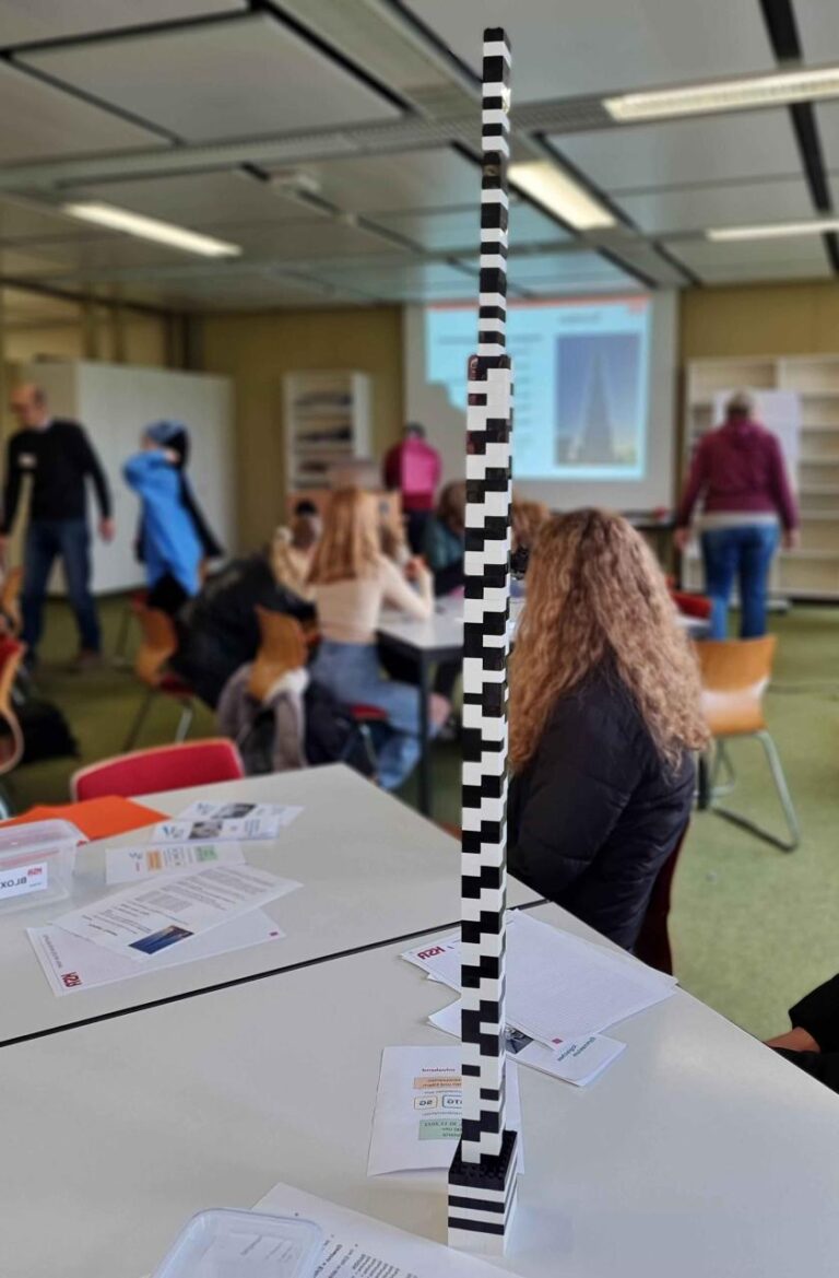 Sehr hoher schmaler Legoturm aus schwarz-weißen Steinchen steht auf einem Tisch. Im Hintergrund arbeiten Schüler zusammen in Gruppen.
